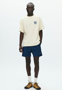 T-shirt couleur crème avec logo bleu, assorti à un short bleu marine, des chaussettes blanches et des chaussures de randonnée marron avec semelles noires. Tenue simple et décontractée.