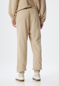 Beige trainingsbroek van zacht materiaal, met een elastische tailleband, geribde manchetten en zijzakken. Draagt met witte sneakers.