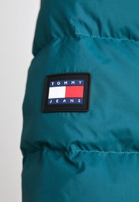 Teal puffer jacket featuring a black rectangular patch with white, red, and blue rectangles labeled "TOMMY JEANS." Smooth, textured fabric.
