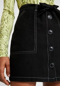 Černá džínová sukně s páskem na zavazování, vyznačující se bílým prošíváním a pěti černými knoflíky, doplněná přední kapsou.