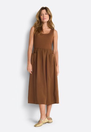 Naine, kellel on pikad lainelised juuksed, kannab varrukateta pruuni midi-kleit taskutega ja beeže lameda tallaga kingi, seisab valge tausta ees.