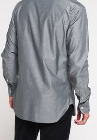 Pánská košile s dlouhým rukávem v jednolité světle šedé barvě. Má zakřivený lem, manžety na knoflíky a design zadního yoku. Hladká textura.