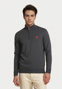 Joven con un suéter de cuello alto gris oscuro con un pequeño logo rojo, combinado con pantalones beige claros, de pie frente a un fondo liso.