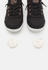 Mustad mustad tennisjalatsid valge tallaga ja roosa kuldse detailiga. Kaasas on kaks rulli valgeid kingapaelu. Suurendatud vaade, keskendudes jalatsitele ja paeltele.
