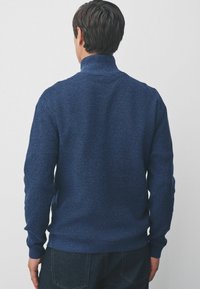 Egy textúrált kék garbót viselő férfi, sötét farmerrel, háttal látható egy világos, egyszínű háttér előtt.