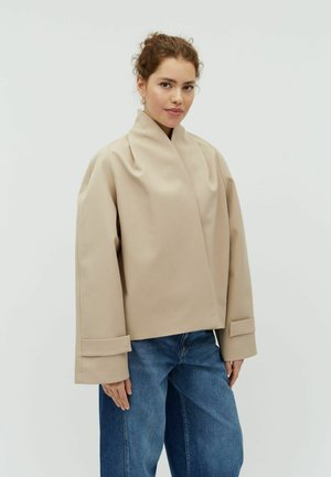 Göndör hajú nő bézs színű, túlméretezett dzsekit és kék farmert visel, világosszürke háttér előtt áll.