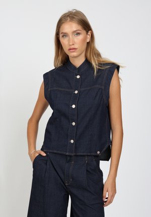 Frau mit langen blonden Haaren, die eine ärmellose dunkelblaue Jeansweste mit silbernen Knöpfen und passende hoch taillierte Hosen vor einem schlichten Hintergrund trägt.