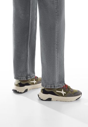 Personne portant un jean gris et des baskets vert olive avec des accents blancs et des lacets rouges, se tenant sur une surface blanche.
