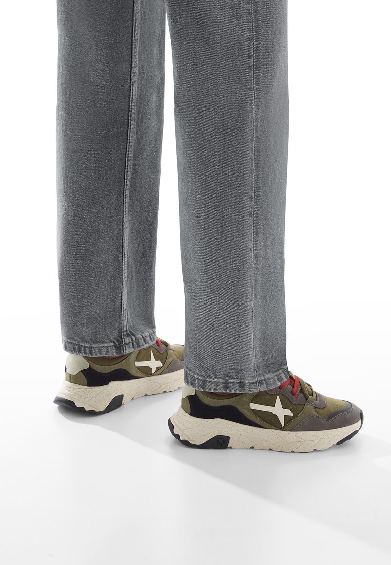 Persoon in grijze jeans en olijfgroene sneakers met witte accenten en rode veters, staande op een witte ondergrond.