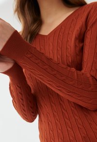 Femme portant un pull en maille torsadée couleur rouille à col en V, ajustant une manche sur un fond uni.