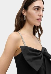 Černé šaty s velkou mašlí v přední části, mají třpytivé ramínka zdobená krystaly a přiléhavý střih.