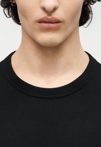 Κοντινό πλάνο του κάτω μέρους του προσώπου και του λαιμού ενός ατόμου που φοράει μαύρο μπλουζάκι με στρογγυλή λαιμόκοψη, με κοντά σγουρά μαλλιά και μικρά σκουλαρίκια σε σχήμα κρίκου.