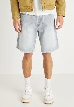 Hellblaue Jeansshorts, knielang mit einem rohen Saum. Getragen mit einem weißen T-Shirt, weißen Socken und beigen Sneakers.
