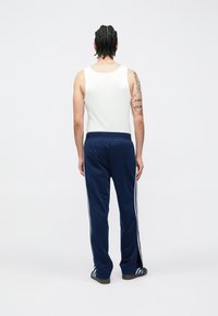 Calças de treino azul-marinho com riscas brancas laterais, cintura elástica e corte ligeiramente afunilado. Usadas com um top de alças branco e calçado preto.