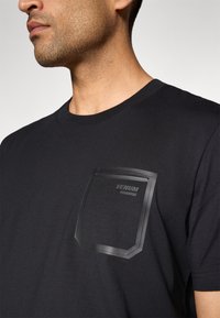 Svart bomull T-skjorte med korte ermer, med en teksturert svart lomme. Lommen har en rektangulær form og preget logo.