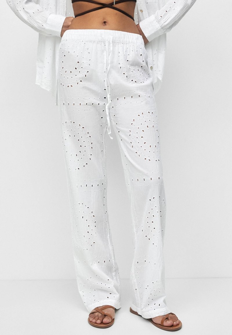 Pantalon blanc fluide avec motifs découpés brodés, taille élastique avec cordon de serrage, et coupe décontractée. Associé à des sandales marron.