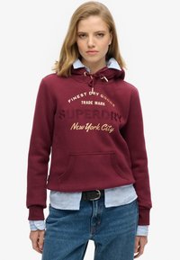 Sudadera burdeos con capucha ajustable, bolsillo frontal y texto impreso. Lleva puesta sobre una camisa de color azul claro, combinada con jeans de mezclilla. Textura de tela suave.