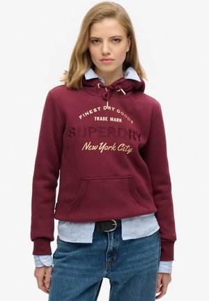 Bordeauxrode hoodie met een trekkoordkap, een borstzak en bedrukt tekst. Draagt over een lichtblauw shirt, gecombineerd met spijkerbroek. Zachte stoftextuur.