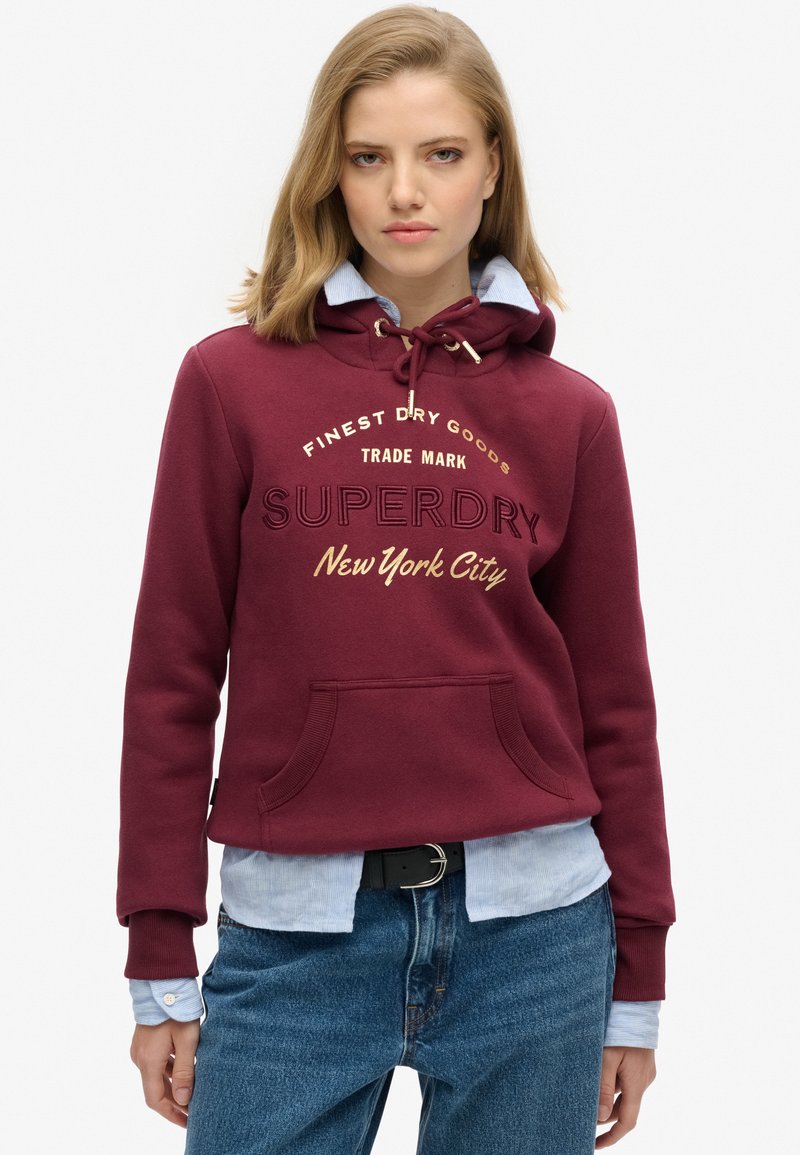 Sudadera burdeos con capucha ajustable, bolsillo frontal y texto impreso. Lleva puesta sobre una camisa de color azul claro, combinada con jeans de mezclilla. Textura de tela suave.