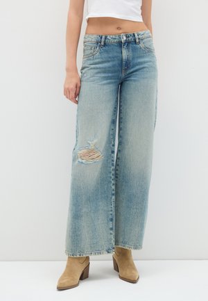 Mujer con vaqueros azules de pierna ancha y cintura alta con una rodilla rasgada y botas de tacón de ante marrón claro sobre un fondo blanco.