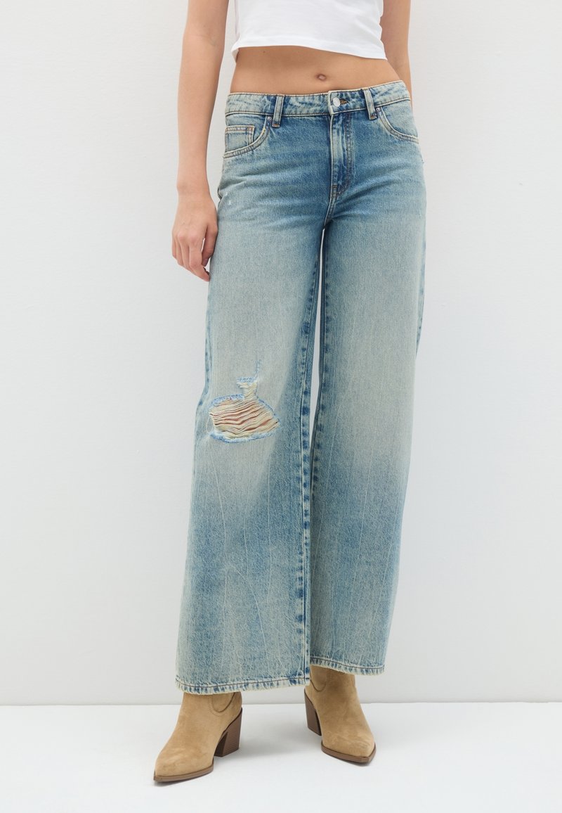 Frau trägt hoch taillierte, weit geschnittene blaue Jeans mit aufgerissenem Knie und hellbraunen Wildleder-Stiefeletten mit Absatz vor weißem Hintergrund.