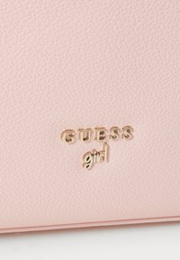 Matériau synthétique texturé rose clair avec un logo "GUESS" embossé en matériel doré et un détail en cursive "girl", présentant une finition lisse.