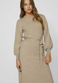Abito a maglia beige con maniche a sbuffo lunghe e vita legata. Presenta un collo rotondo e una consistenza liscia, raggiungendo la lunghezza a metà polpaccio.