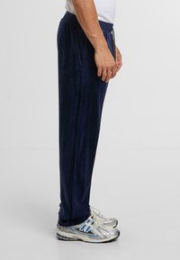 Mężczyzna w granatowych aksamitnych spodniach i srebrnych butach do biegania, stojący bokiem na jednolitym jasnym tle.