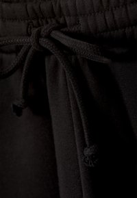 Un pantalon de survêtement noir présente un cordon tissé avec deux extrémités nouées, une taille élastique et une texture lisse semblable au coton.