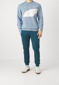 Sudadera azul clara con acentos blancos y pantalones jogger en tonos verde azulado. Presenta puños de canalé y una cintura elástica. Zapatillas blancas con acentos beige.