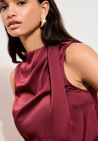 Bordeauxrotes ärmelloses Oberteil aus glattem, glänzendem Stoff mit drapierten Details an der Schulter. Goldene Ohrringe akzentuieren den Look.