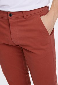 Rostfärgade bomullsbyxor med rak passform, sidofickor och en enda knappstängning vid midjan. Slät textur och minimal sömnad.