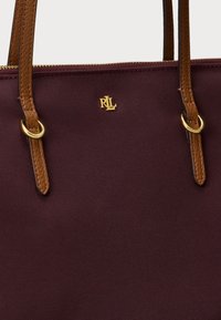 Bordová taška cez plece s hnedými koženými rúčkami a zlatými kovovými doplnkami. Obsahuje textúrovanú tkaninu a razený logo na prednej strane.