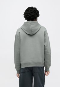 Personne aux cheveux bouclés foncés portant un sweat à capuche gris clair et un pantalon foncé, debout dos à un fond blanc uni.