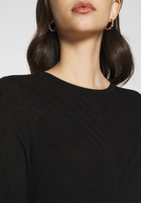 Schwarzes strukturiertes Strickoberteil mit Rundhalsausschnitt und dezenten Chevron-Mustern, die diagonal über den Stoff verlaufen. Goldene Creolen sind sichtbar.