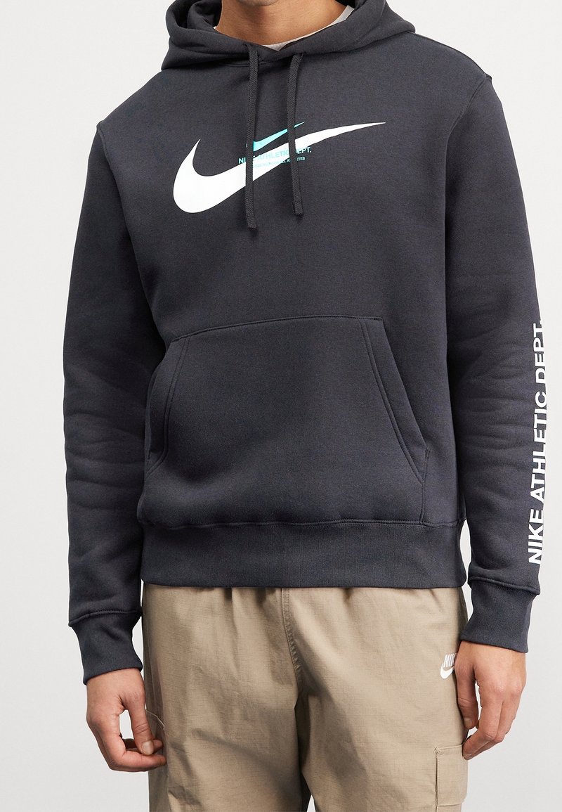 Hoodie Nike gris foncé avec un grand logo blanc, poche kangourou et texte sur la manche. Fabriqué en tissu doux avec une capuche et des cordons de serrage.