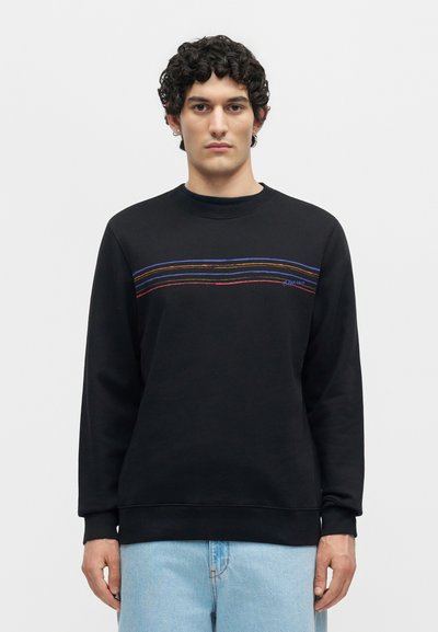 Jeune homme aux cheveux bouclés portant un sweat-shirt noir avec des bandes horizontales multicolores sur la poitrine et un jean bleu clair.