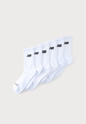 Seks par hvide crew-sokker med sorte New Balance-logoer nær toppen, arrangeret overlappende på en ensartet lys baggrund.