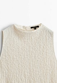 Haut sans manches crème texturé avec col montant et fermeture éclair arrière, posé à plat sur un fond blanc.