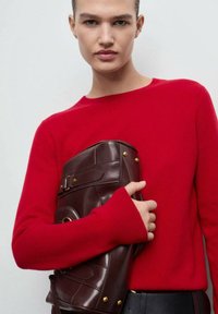 Czerwony sweter dzianinowy z wydłużonymi rękawami; model trzyma ciemnobrązową skórzaną torbę z złotymi detalami i zapięciem.