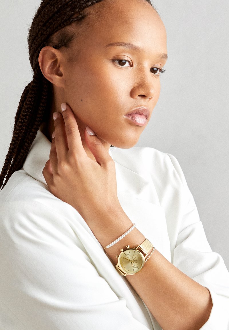 Goldene Armbanduhr mit rundem Ziffernblatt, Mesh-Band und drei Subdials. Begleitet von einem zarten Perlenarmband. Weiße Kleidung ergänzt die Accessoires.