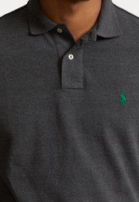Mörkgrå polo skjorta i texturerad bomull, med klassisk krage, två knappar och en liten grön logotyp på vänster bröst.