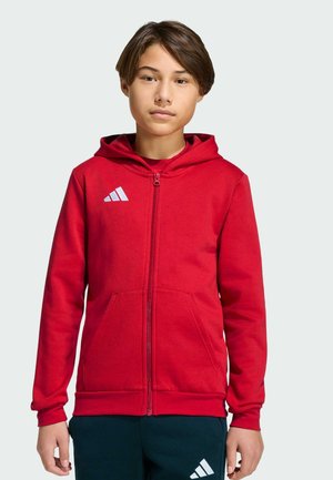 Jeune personne portant un sweat à capuche rouge zippé avec un logo blanc Adidas et un pantalon foncé avec un logo assorti, debout devant un fond uni.