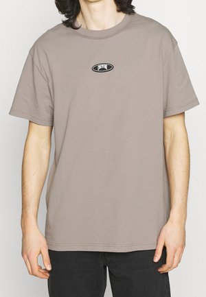 Mand iført en taupe kortærmet T-shirt med et lille ovalt "COUTURE" logo på brystet og sorte bukser.