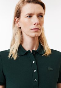 Polo shirt verde scuro realizzato in tessuto testurizzato, con colletto, cinque bottoni e un piccolo logo ricamato sul lato sinistro del petto.