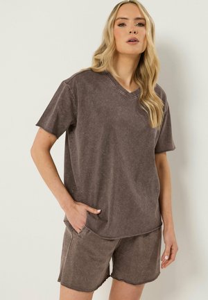 Lyshåret kvinde iført løs brun vasket V-hals T-shirt og matchende shorts med den ene hånd i lommen, stående mod en ensfarvet baggrund.