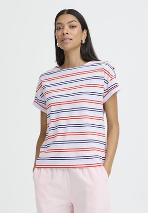 Femme aux longs cheveux foncés portant des boucles d'oreilles en argent en forme d’anneaux, un T-shirt blanc à rayures horizontales rouges et bleu marine, et un pantalon rose clair, les mains dans les poches.