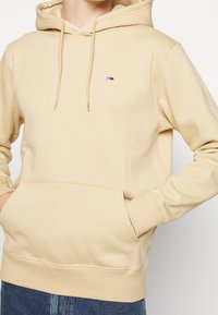 Sudadera con capucha beige que incluye un bolsillo canguro, capucha con cordón ajustable, puños acanalados y un pequeño logotipo bordado en el pecho. Tela suave y lisa.