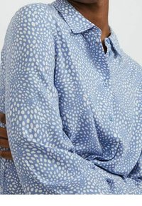 Ljust blå skjorta med knappar och ett prickigt mönster i olika storlekar, med krage och långa ärmar i en mjuk tygkvalitet.