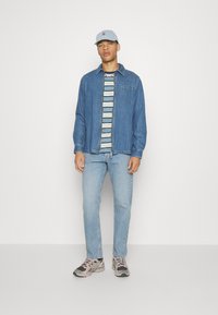 Giubbotto in denim blu, t-shirt a righe sotto, jeans azzurri chiari e sneakers multicolori. Berretto azzurro chiaro con dettagli del logo.